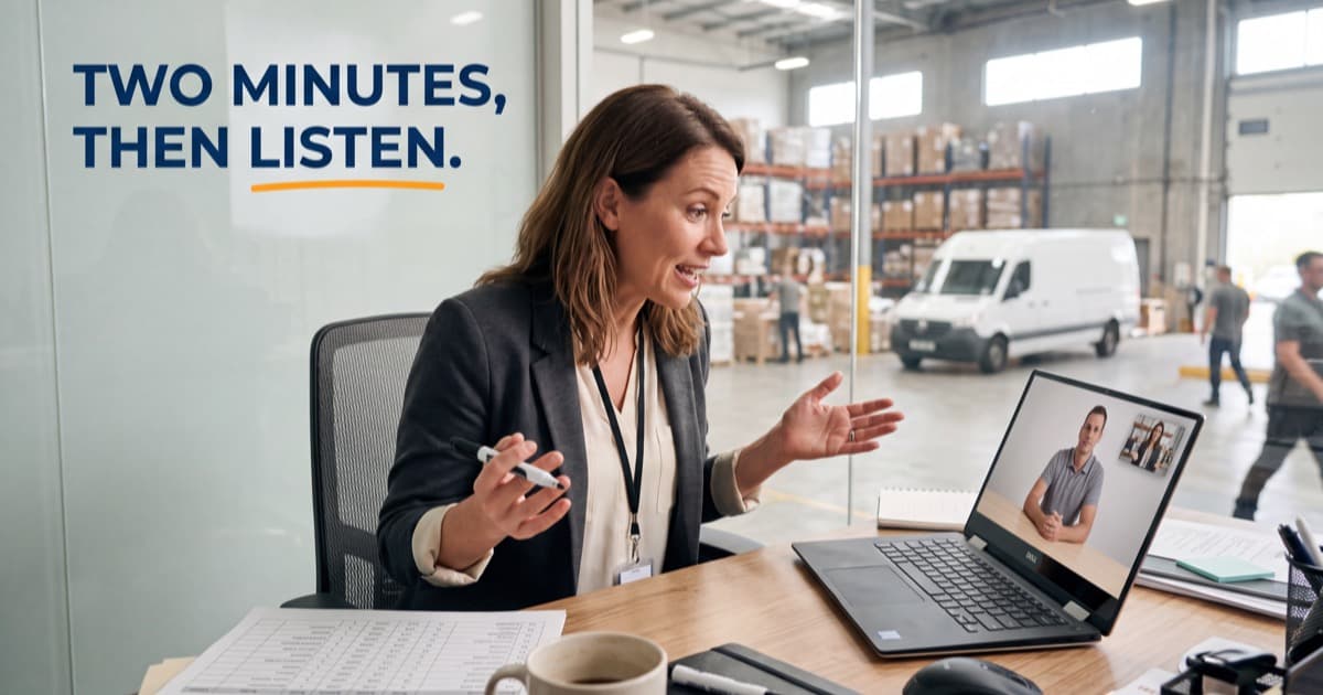 A saleswoman in a warehouse office gestures mid-sentence during a laptop video call with a buyer, printed reports and a mug spread across her bright desk